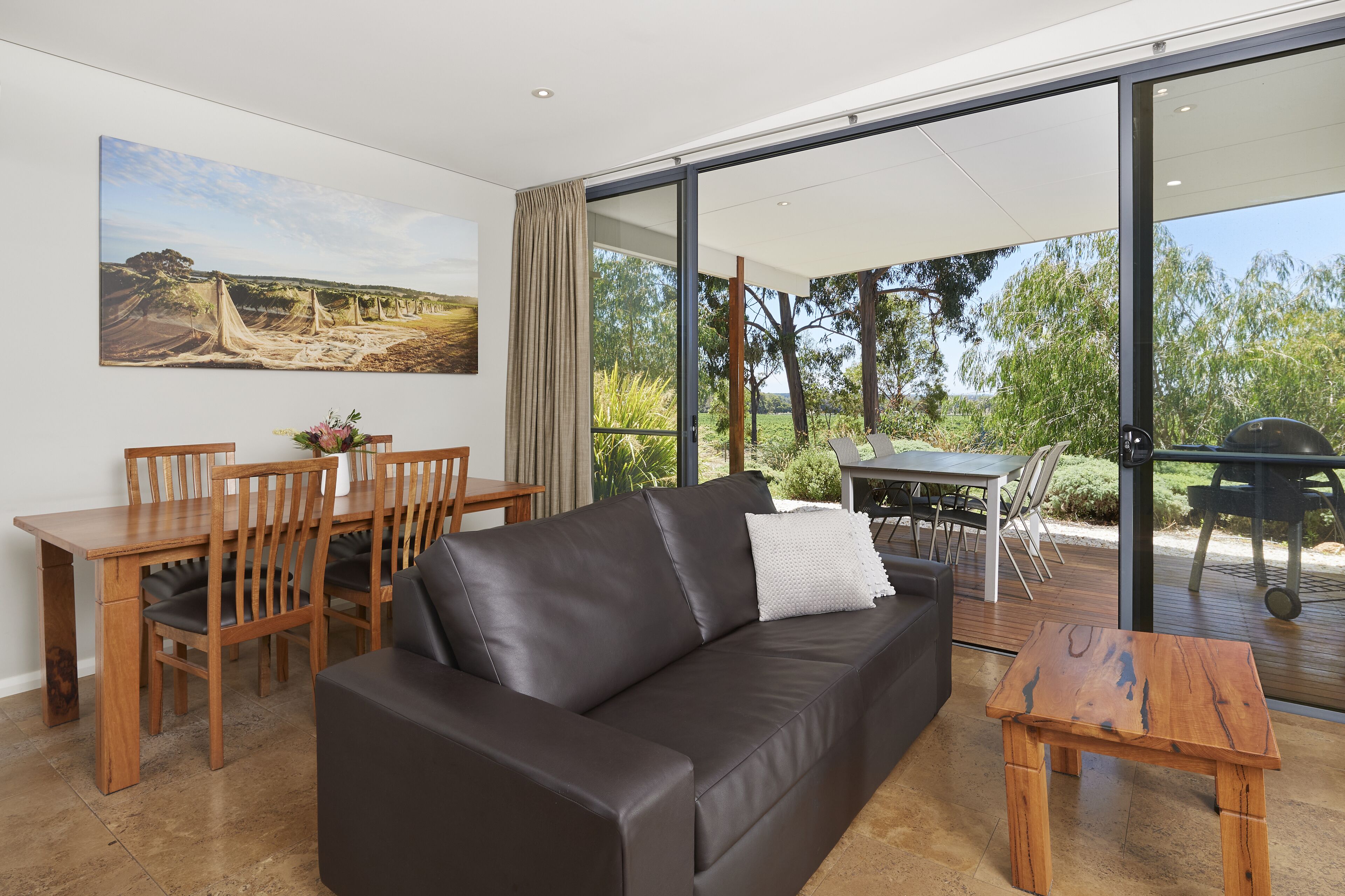 A modern living room with a leather sofa, wooden dining table, and chairs. There is a large window with a view of greenery and a painting on the wall.