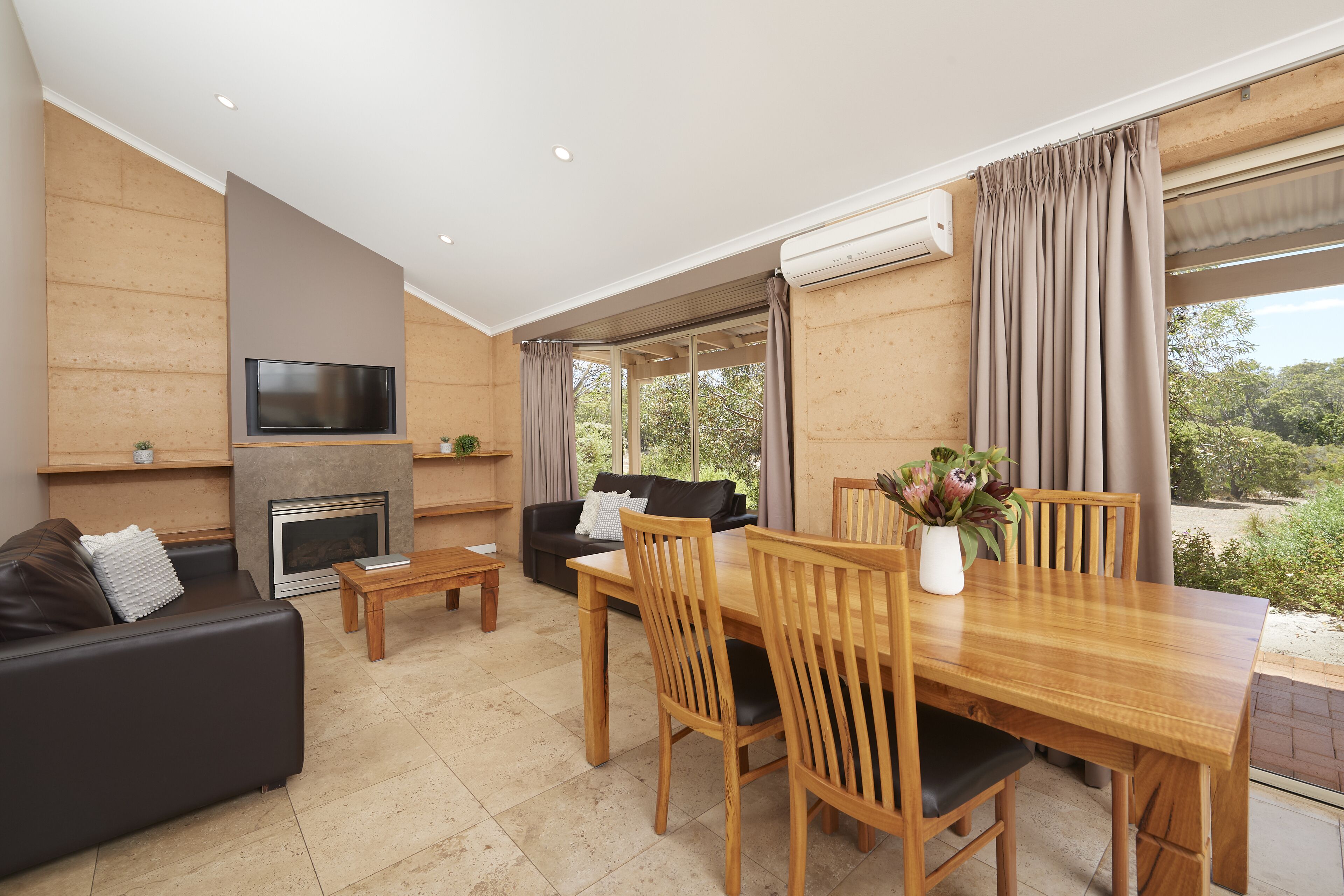 A modern living room with a fireplace, a TV mounted on the wall, a dining area with wooden furniture, and large windows offering a view of greenery outside.