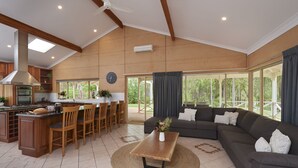 A spacious living area with a large sofa, a coffee table, and a dining area with chairs and a kitchen in the background. - Eight Willows Retreat (Metricup)