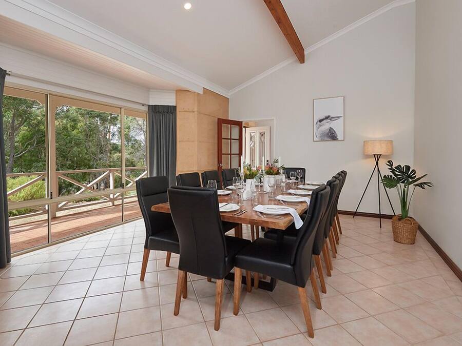 Un comedor con una mesa grande dispuesta para seis personas, rodeado de sillas negras, ventanas amplias con cortinas y vistas al exterior con vegetación.