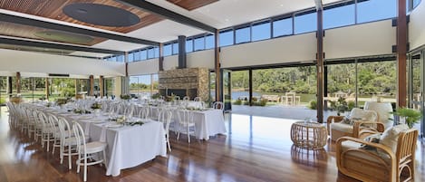 Un comedor interior amplio con largas mesas dispuestas para una comida, grandes ventanales que ofrecen vistas al exterior y un diseño de techo moderno.