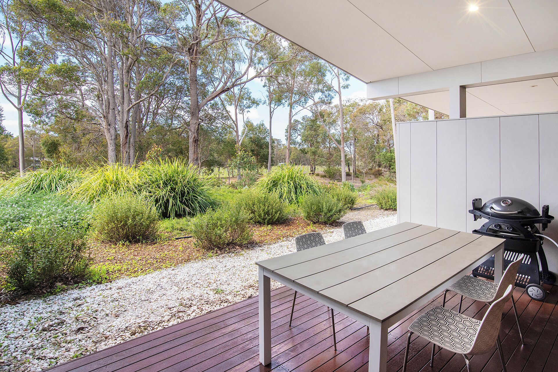 Un espacio exterior para comer con mesa y sillas, una parrilla y vistas a un paisaje natural con árboles y arbustos.
