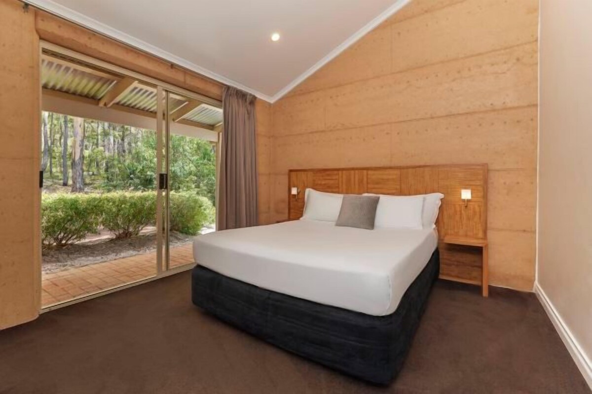 A modern bedroom with a large bed, wooden walls, and a glass door leading to a garden.