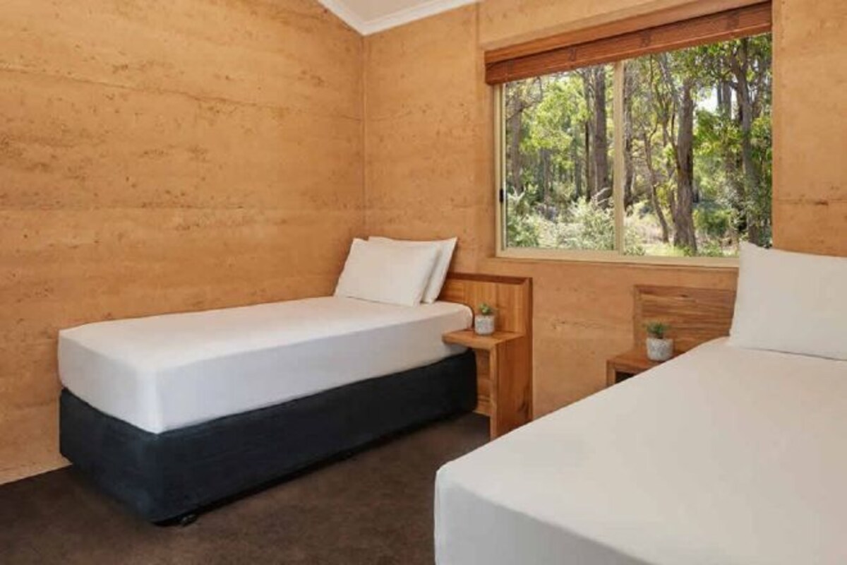 A bedroom with two beds, wooden walls, a window with a view of trees, and a small bedside table with a plant.
