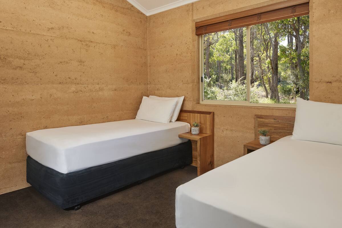 Un dormitorio con dos camas, una ventana con vistas al bosque y una mesita de noche con una planta.