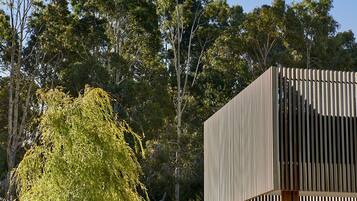 Un edificio moderno con fachada de madera y base de piedra, rodeado por un jardín ajardinado con diversos arbustos y un árbol grande.
