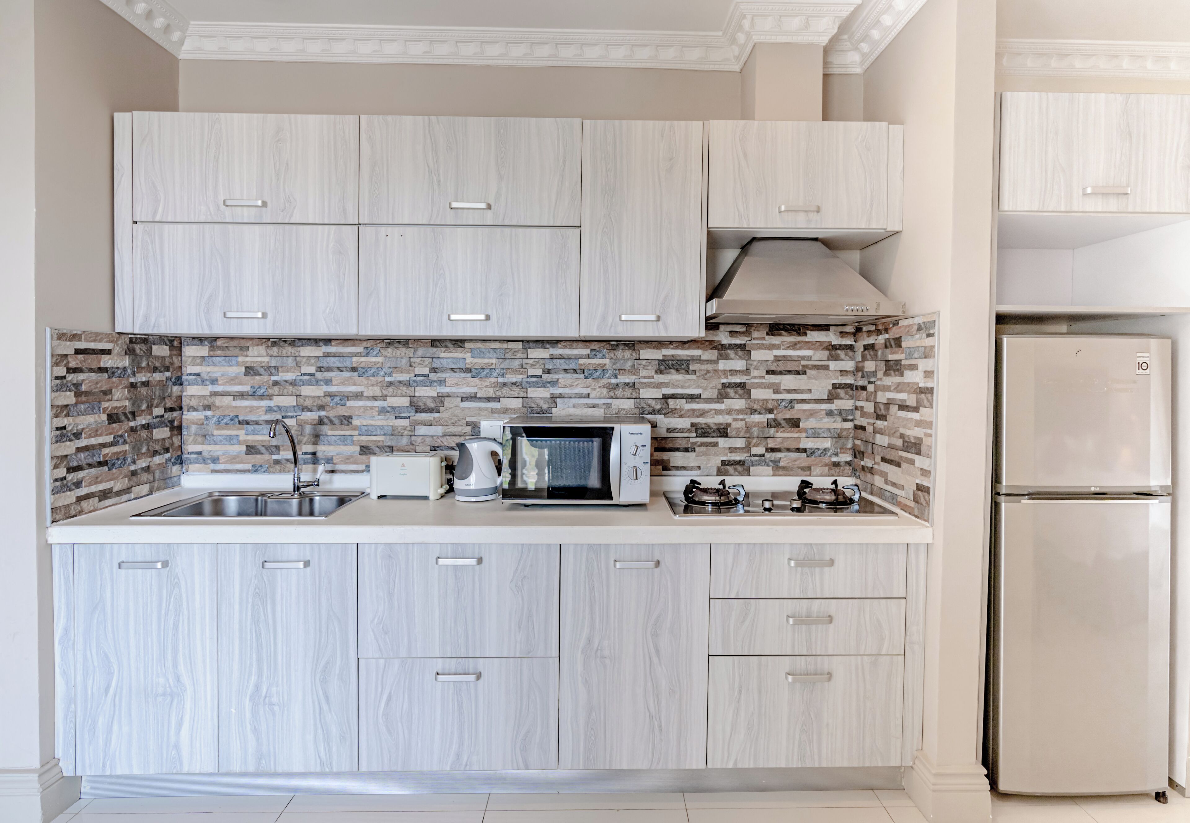 A kitchen with light wood cabinets, a tiled backsplash, a microwave, a kettle, and a refrigerator.