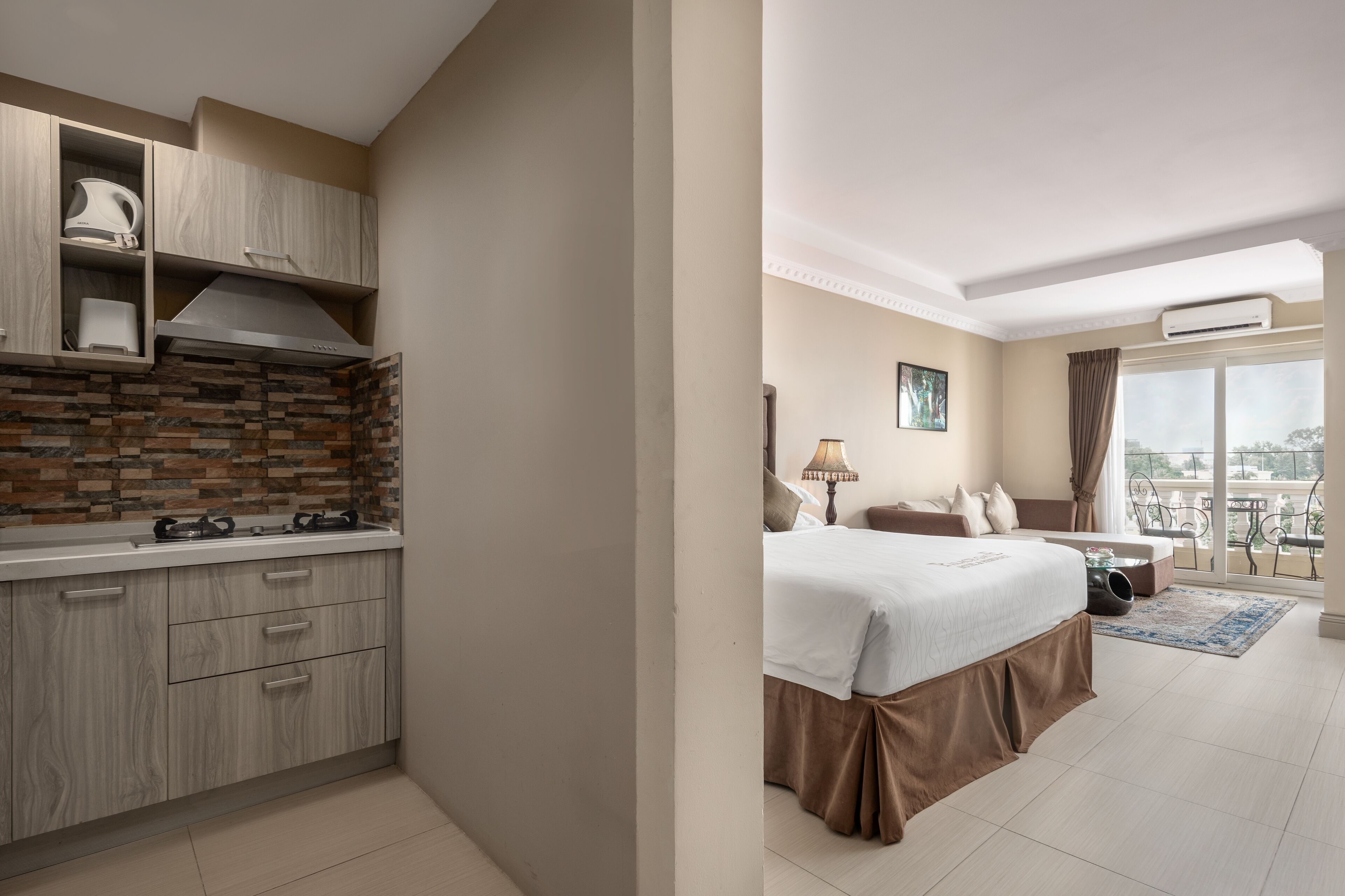A modern hotel room with a kitchenette, a large bed, a balcony with a view, and a window with curtains.