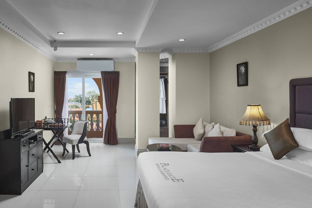 A hotel room with a bed, a desk with a computer, a chair, a sofa, and a balcony with curtains.