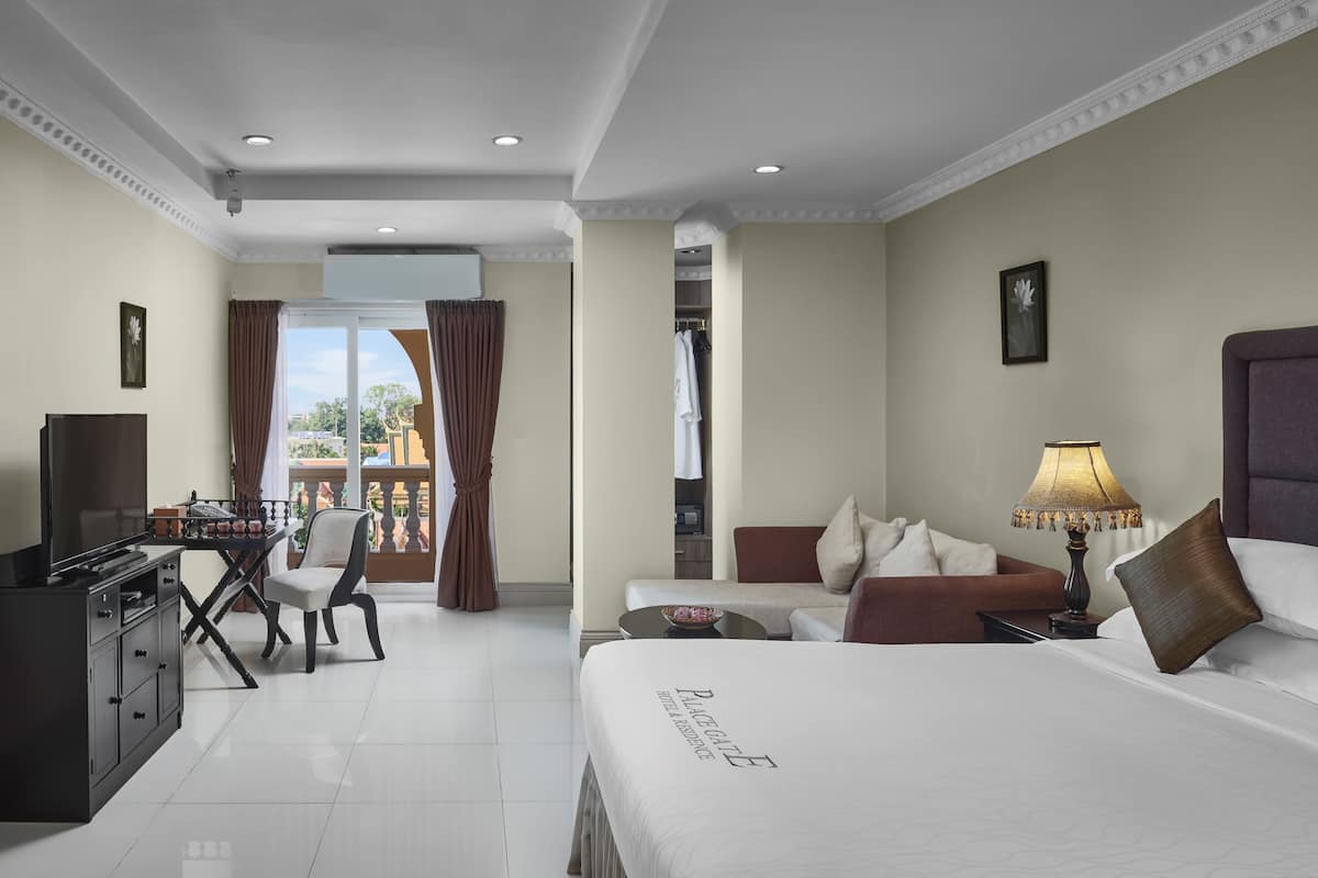 A hotel room with a bed, a desk with a TV, a chair, a sofa, and a balcony with curtains.