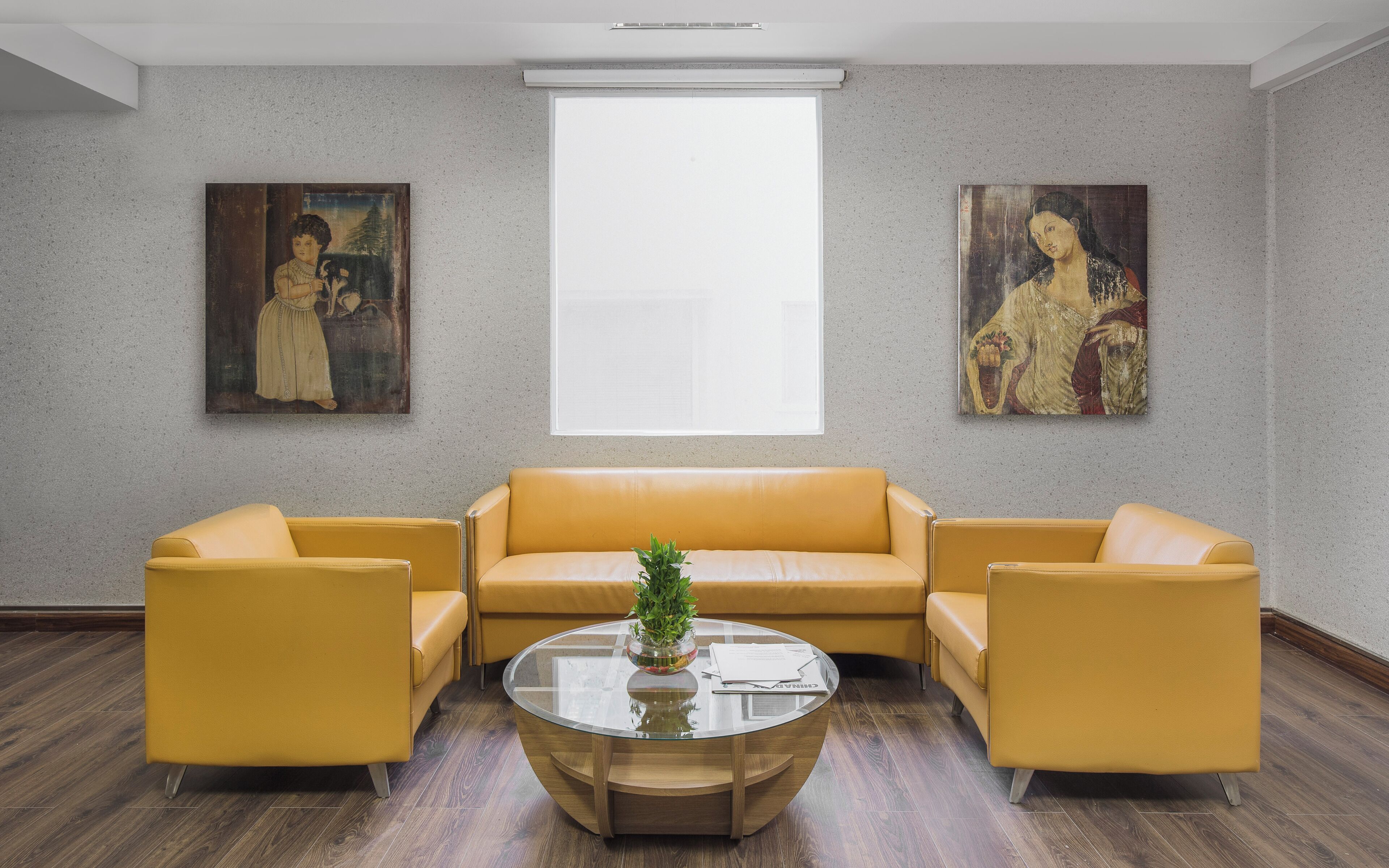 A room with a yellow sofa set, a glass coffee table with a plant, and two paintings on the wall.