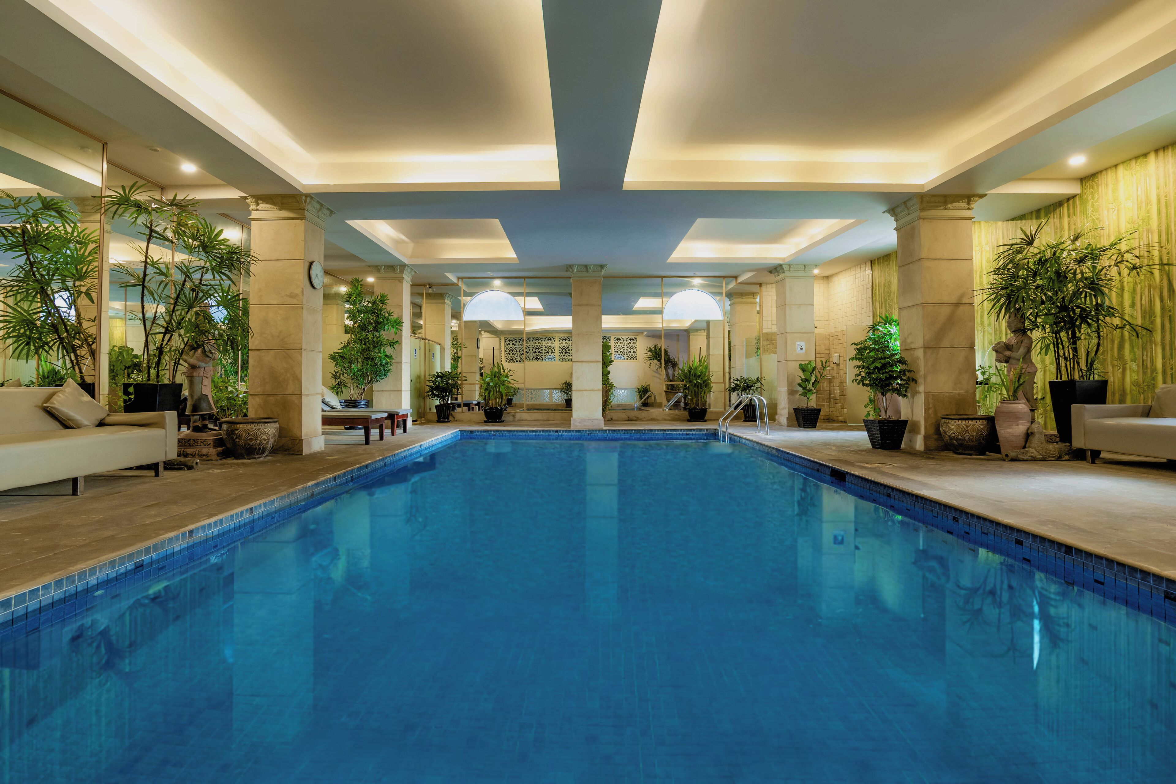 Una amplia piscina interior con suelo de baldosas, rodeada de plantas en macetas y un sofá.