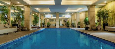 A large indoor swimming pool with a tiled floor, surrounded by potted plants and a sofa.