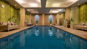 A large indoor swimming pool with a tiled floor, surrounded by beige sofas and potted plants. - Palace Gate Hotel & Residence by EHM (Phnom Penh)