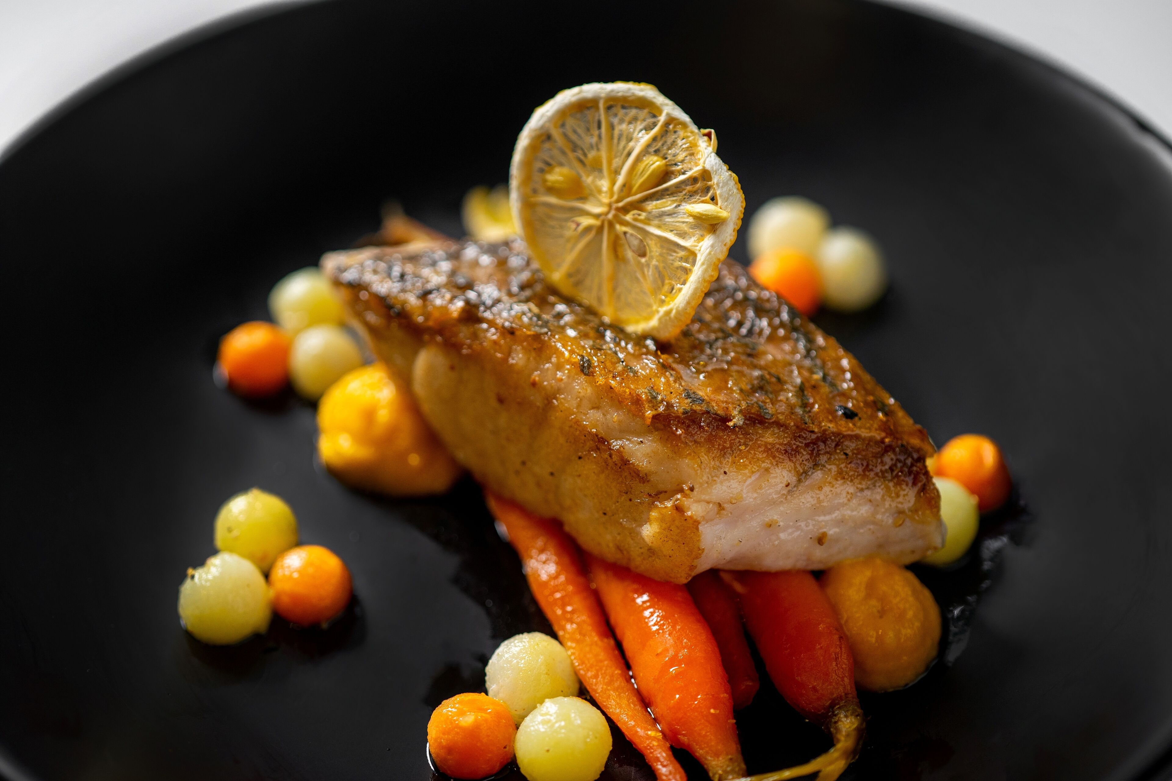 A dish with a fish fillet, lemon slice, and small round vegetables on a black plate.