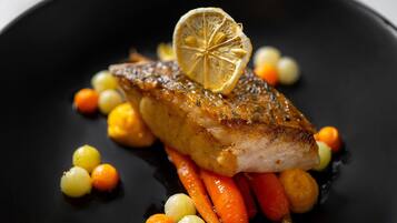 A dish with a fish fillet, lemon slice, and small round vegetables on a black plate.