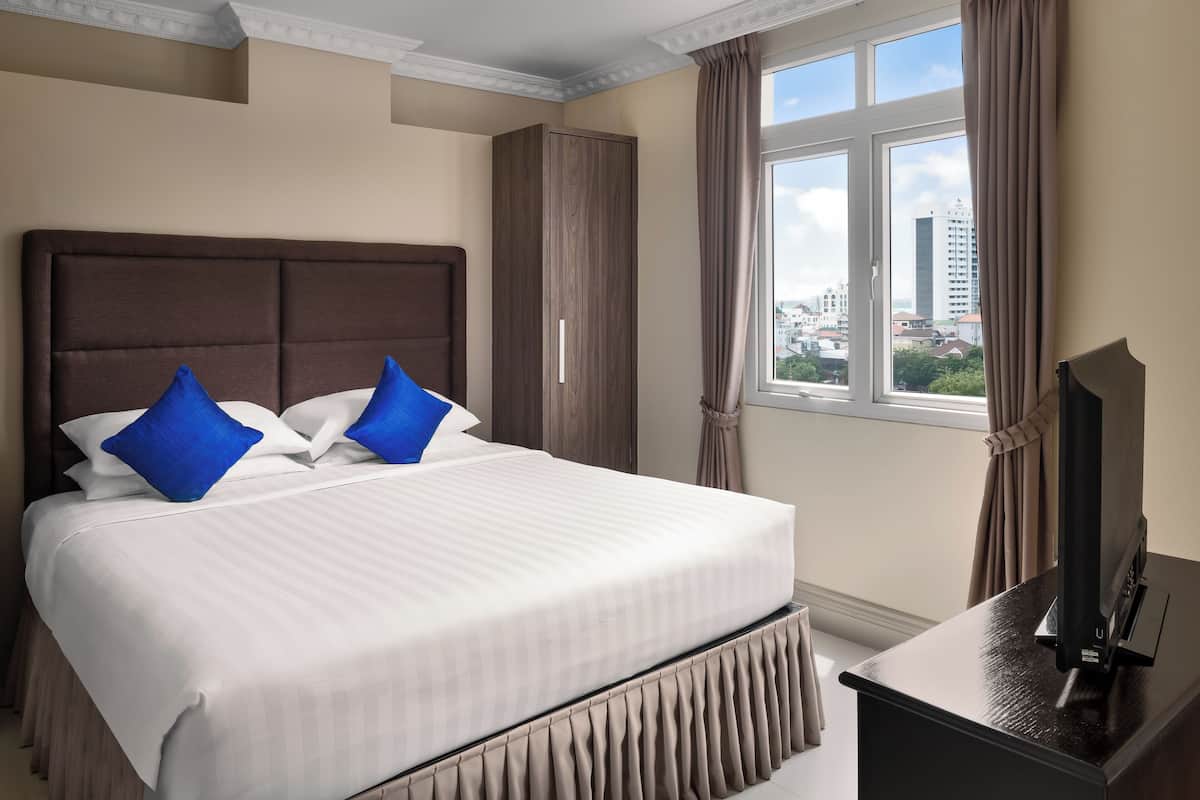 A neatly made bed with white and blue pillows, a dark headboard, and a view of the city through a window with curtains.