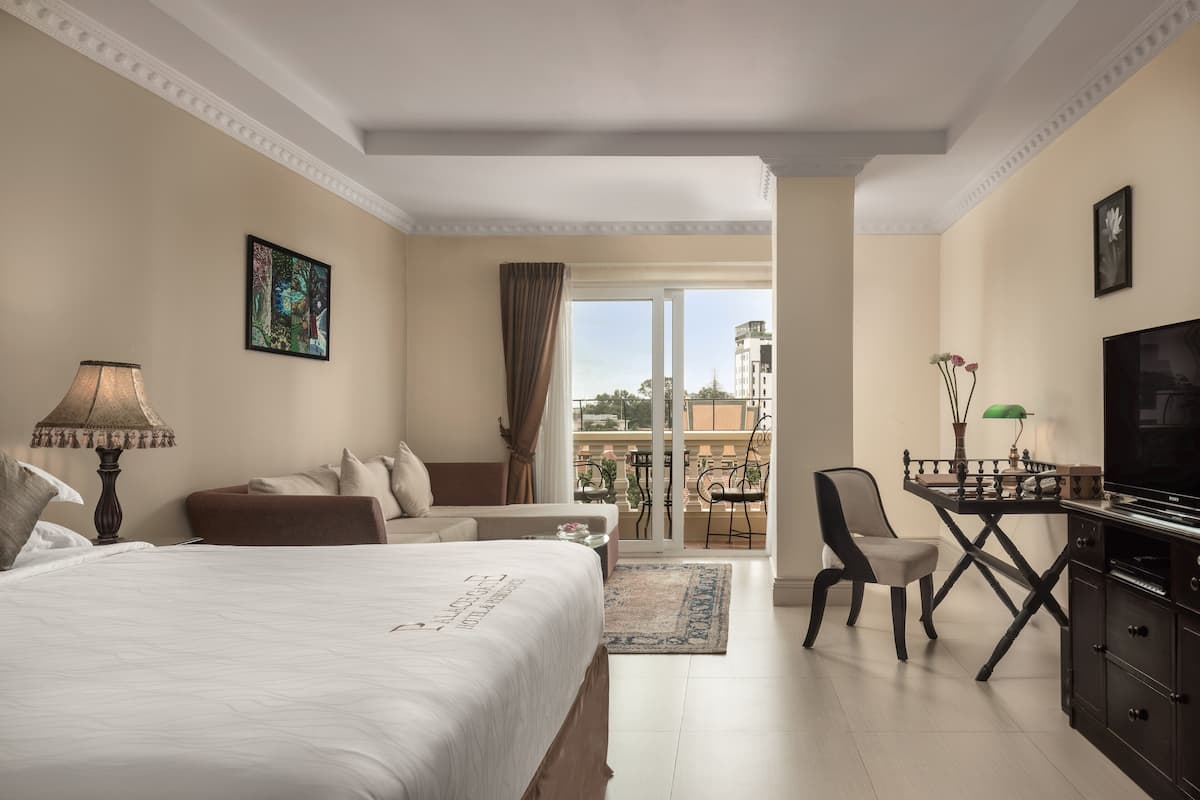 A hotel room with a large bed, a desk with a chair, a television, and a balcony with a view of buildings.