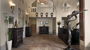 A grand hallway with a high ceiling, ornate wall sconces, and a large chandelier.