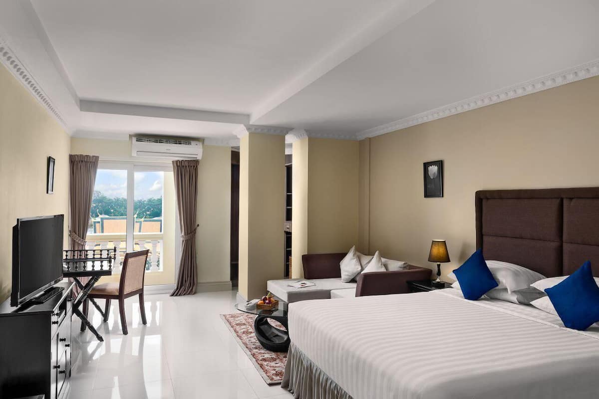 A hotel room with a bed, a desk with a TV, a sofa, a lamp, and a view of the outdoors.