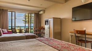A hotel room with two beds, a desk, a chair, a TV, and a balcony with a view of the sea.