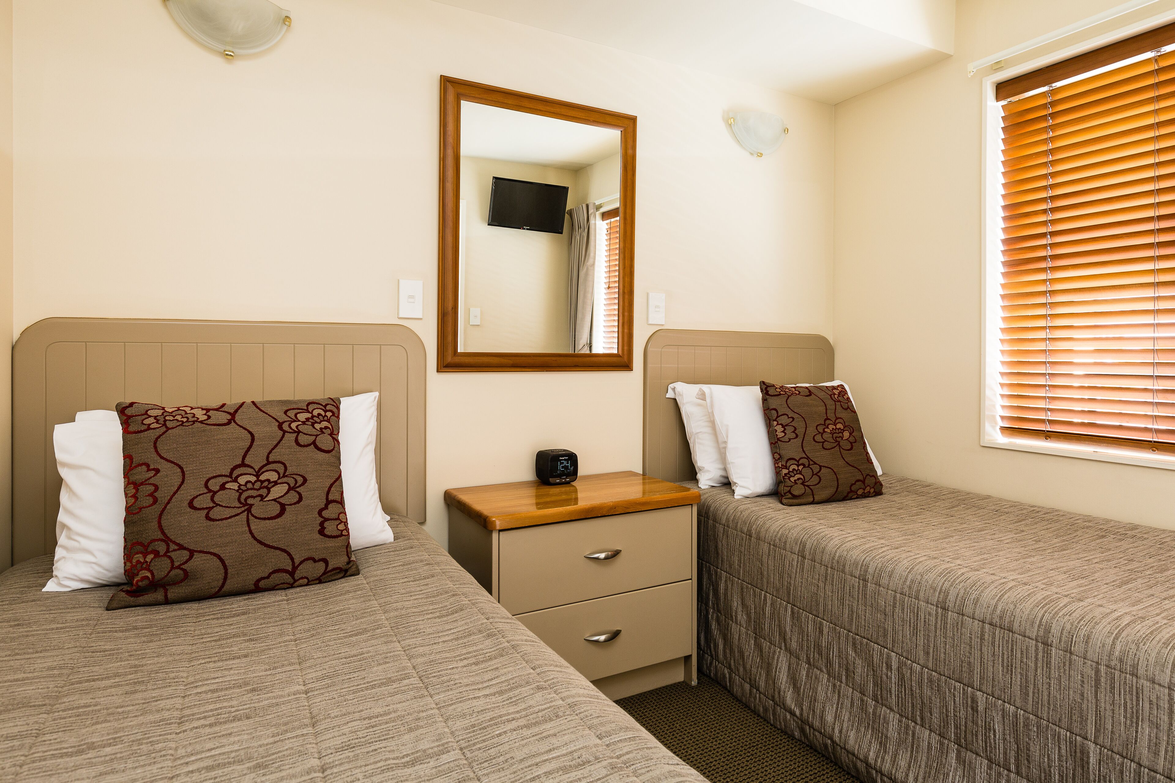 Une chambre avec deux lits simples, une table de chevet avec une horloge, un miroir et une fenêtre avec des stores.