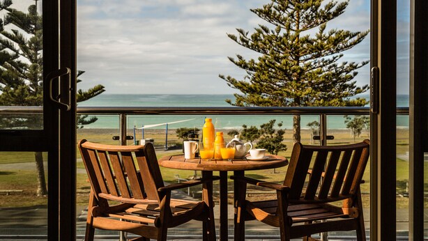 A wooden table with two chairs and a breakfast setup on a balcony overlooking a scenic view of the ocean and trees.