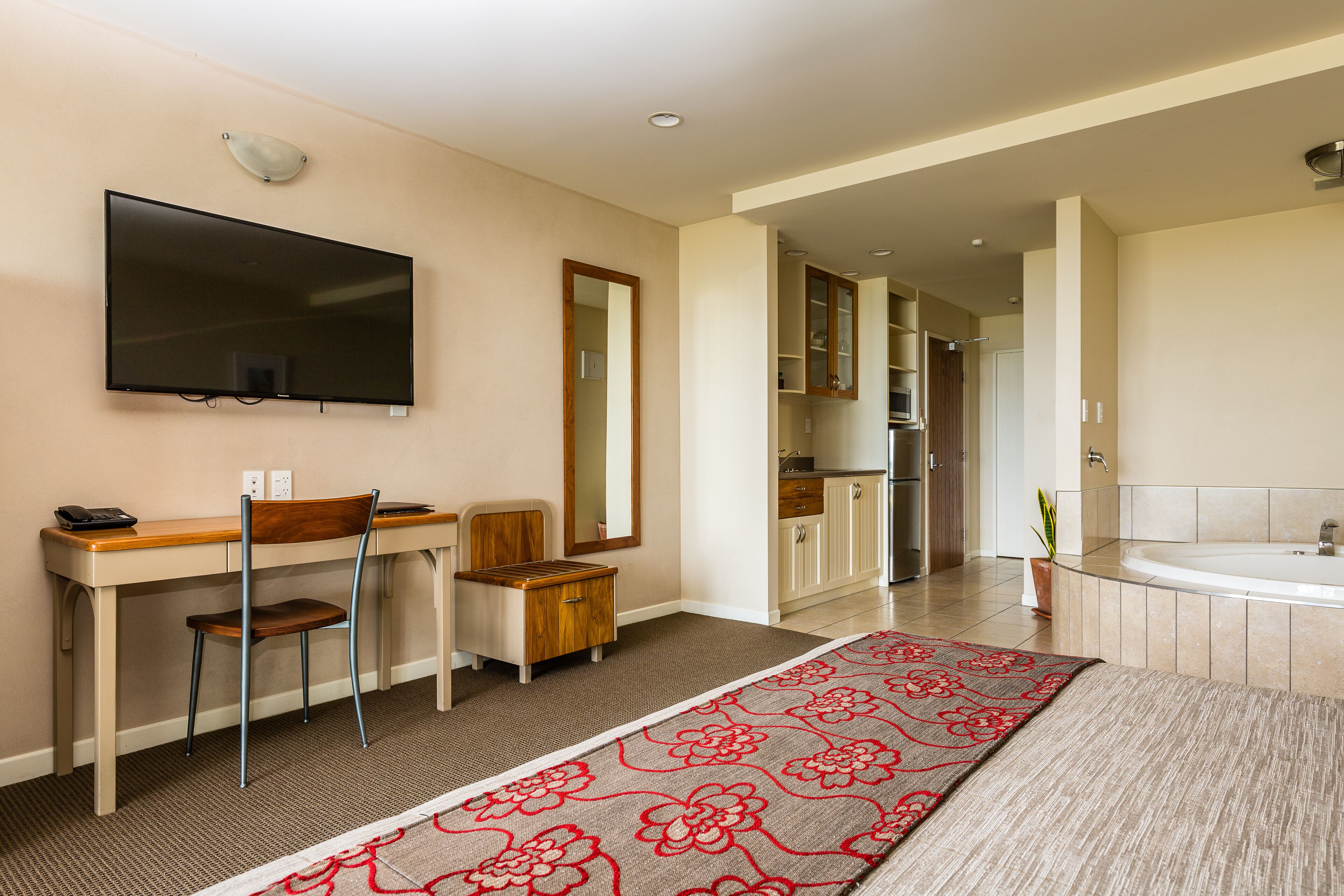 A modern hotel room with a flat-screen TV, a desk with a chair, a large mirror, a bathroom with a bathtub, and a kitchen area.