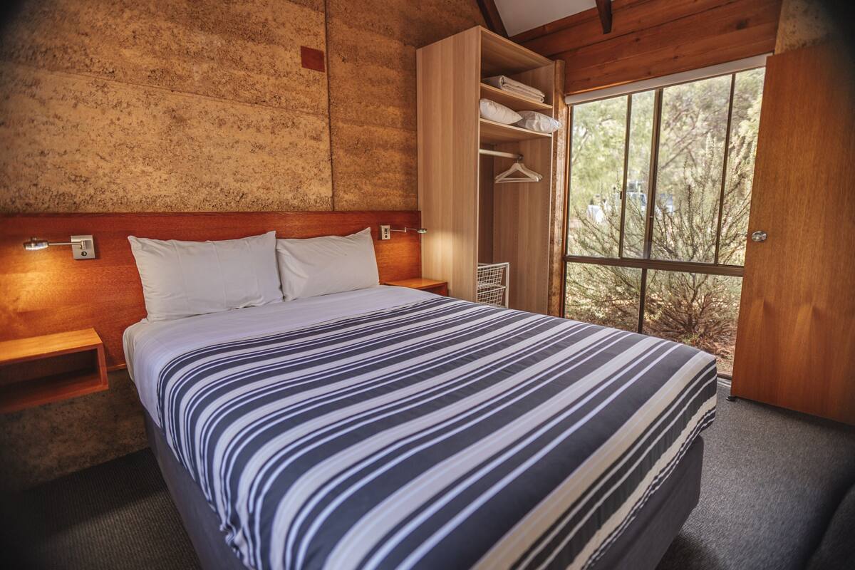 A bedroom with a bed, wooden headboard, a nightstand, a wardrobe, and a window with a view of trees.