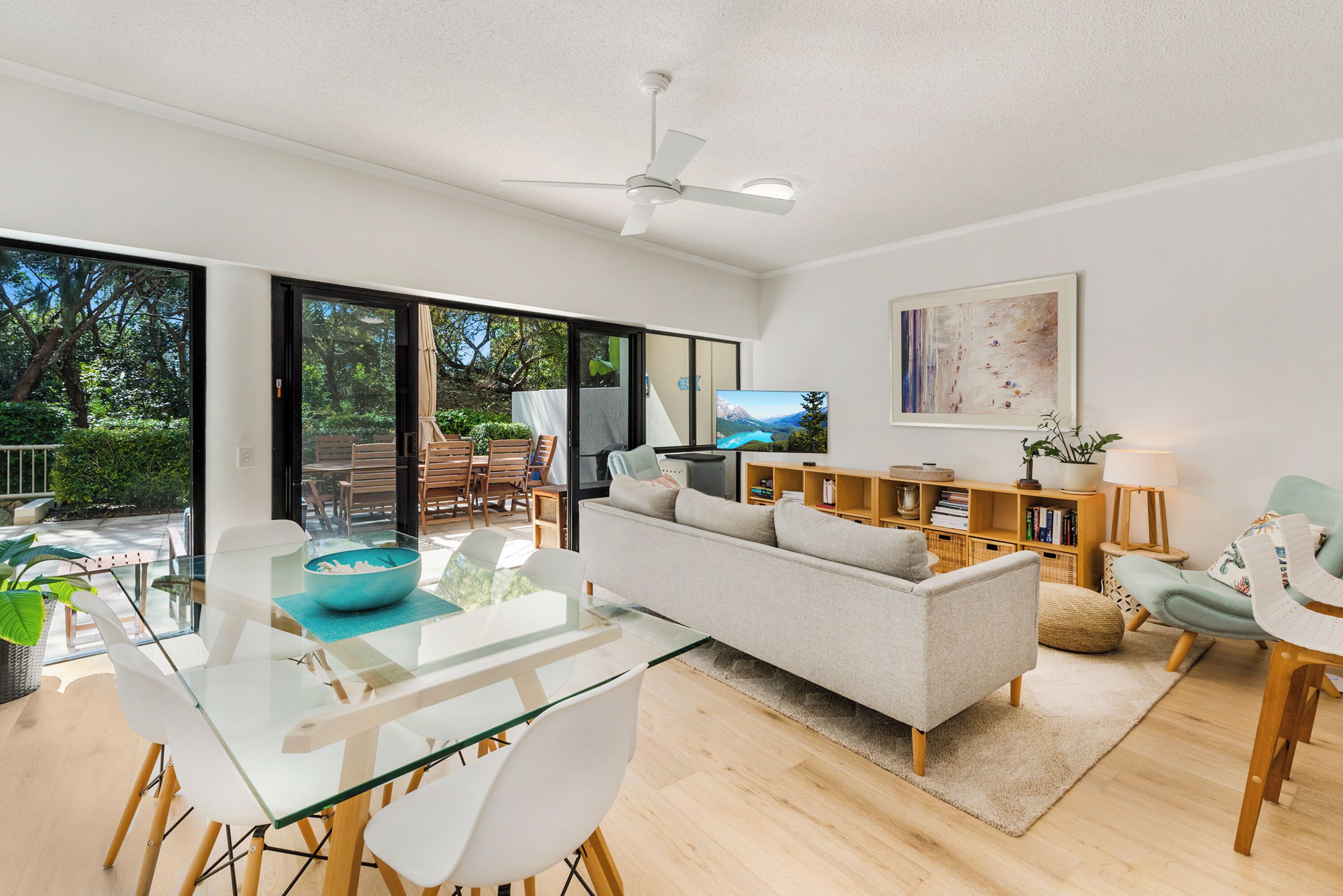 A modern living room with a glass dining table, a sofa, and a ceiling fan.