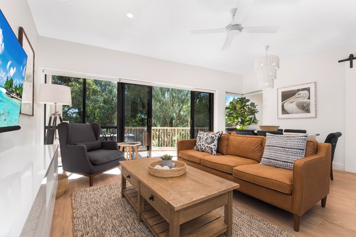 A modern living room with a sofa, armchair, coffee table, and a large window offering a view of greenery.