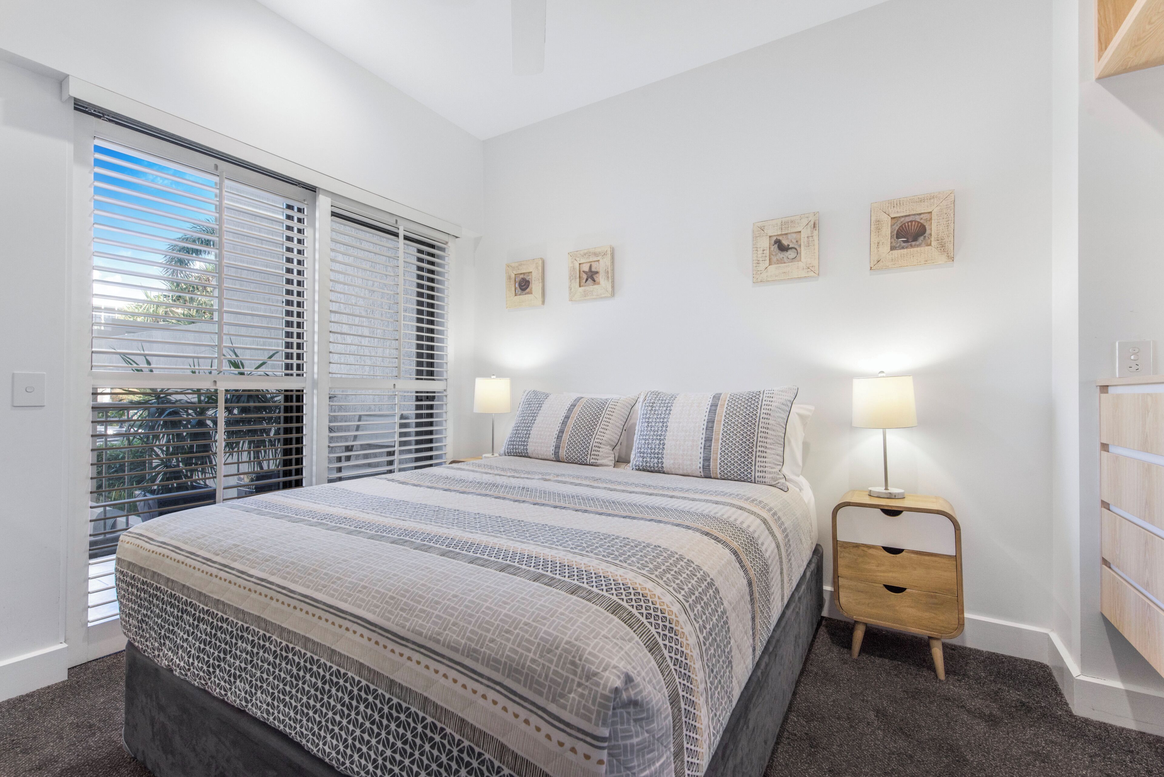 A bedroom with a large bed, a nightstand, a window with blinds, and wall art.