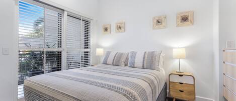 A bedroom with a large bed, a nightstand, a window with blinds, and wall art.