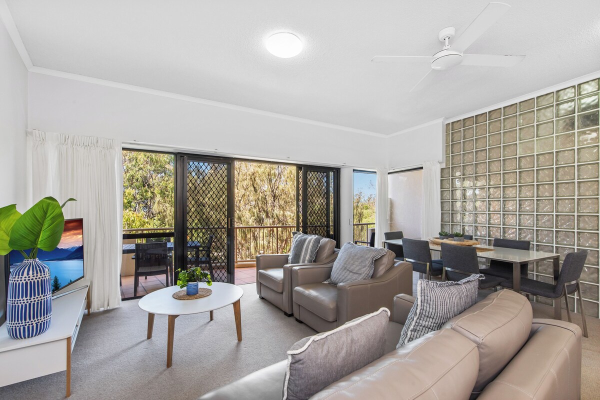 A modern living room with a sofa, coffee table, and TV, connected to a balcony with a dining area.