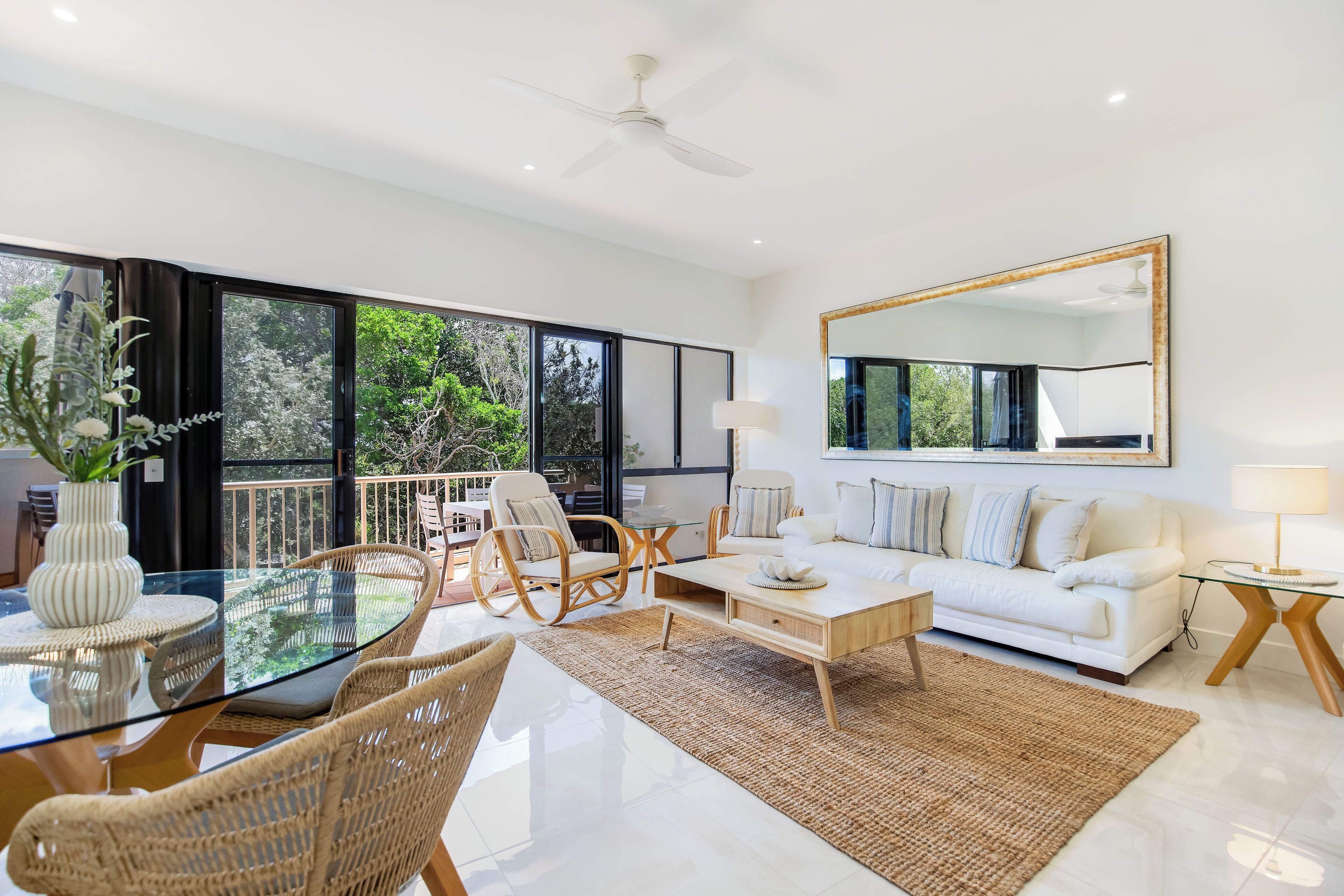 A modern living room with a glass dining table, wicker chairs, a white sofa, and a large mirror.