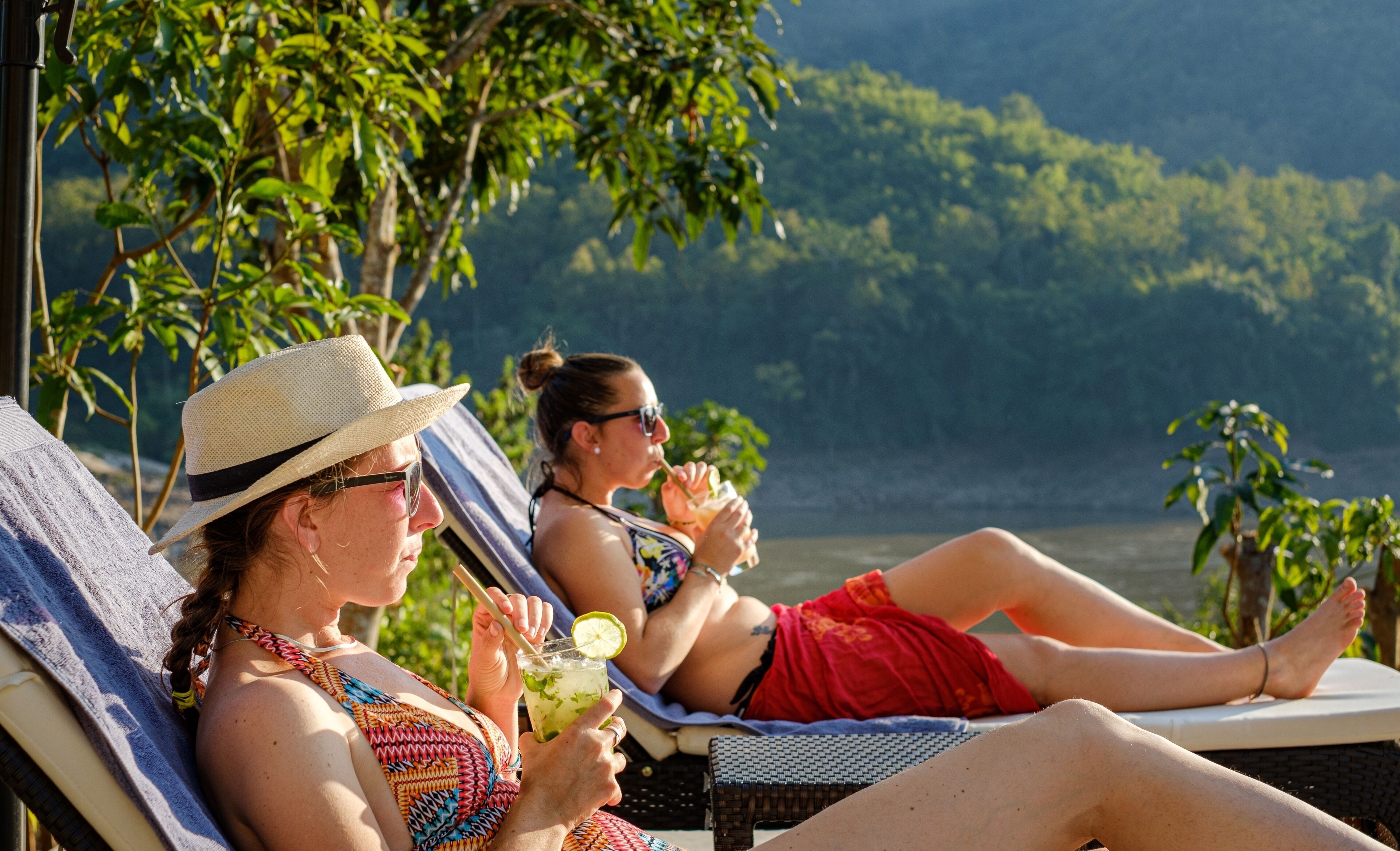 Duas pessoas relaxando em espreguiçadeiras perto da água, com vegetação e colinas ao fundo.