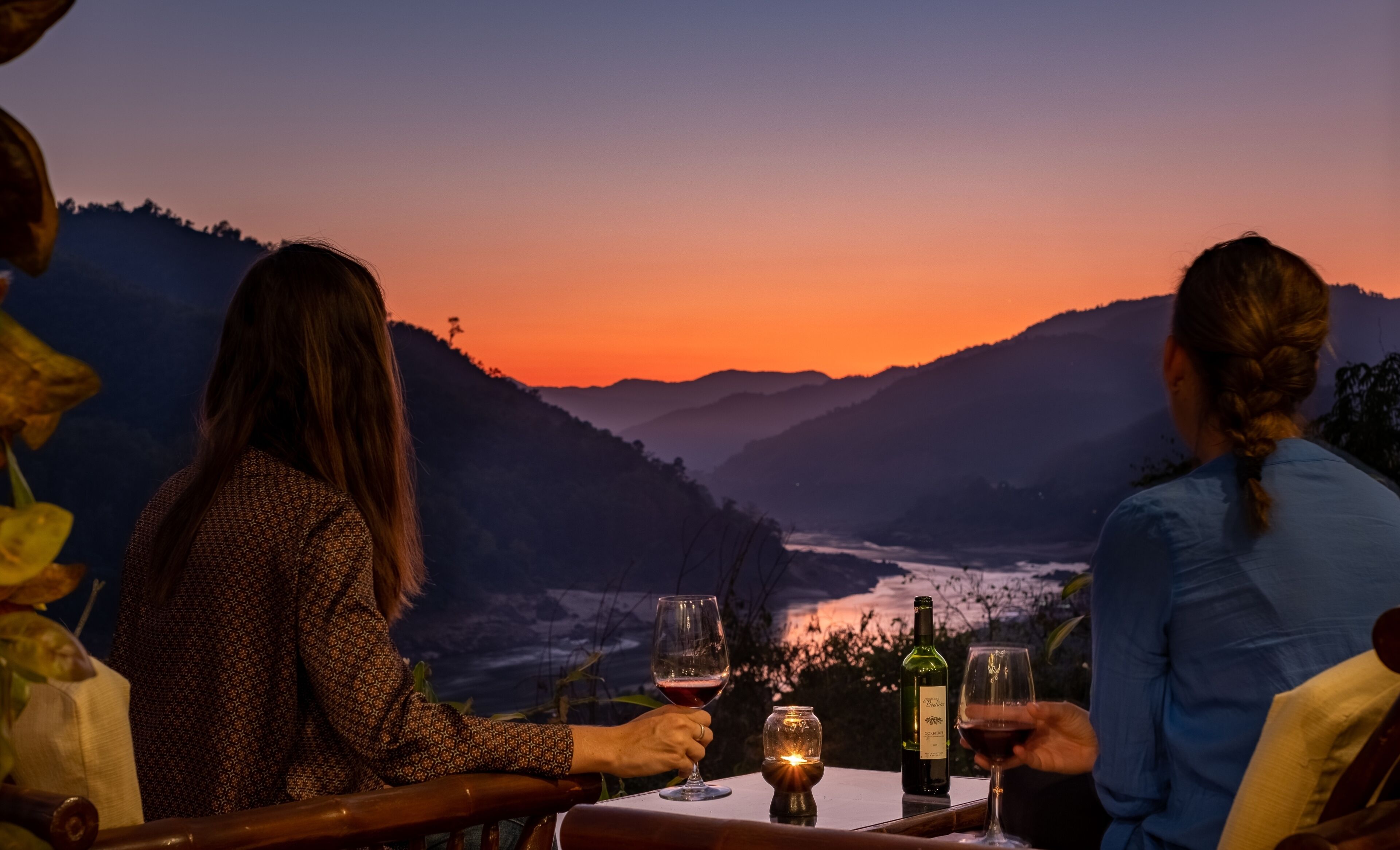 A table with a bottle of wine and two glasses, set against a scenic mountain backdrop during sunset.