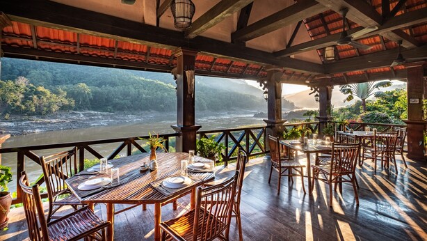 A restaurant with wooden tables and chairs, a view of a river, and a balcony with plants. - Sanctuary Pakbeng Lodge (Pakbeng)
