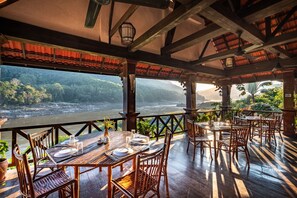 Un restaurante con mesas y sillas de madera, vista al río y un balcón con plantas.