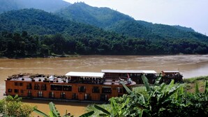 A riverboat with multiple decks and a covered upper deck, situated on a river with a forested hillside in the background. - Sanctuary Pakbeng Lodge (Pakbeng)