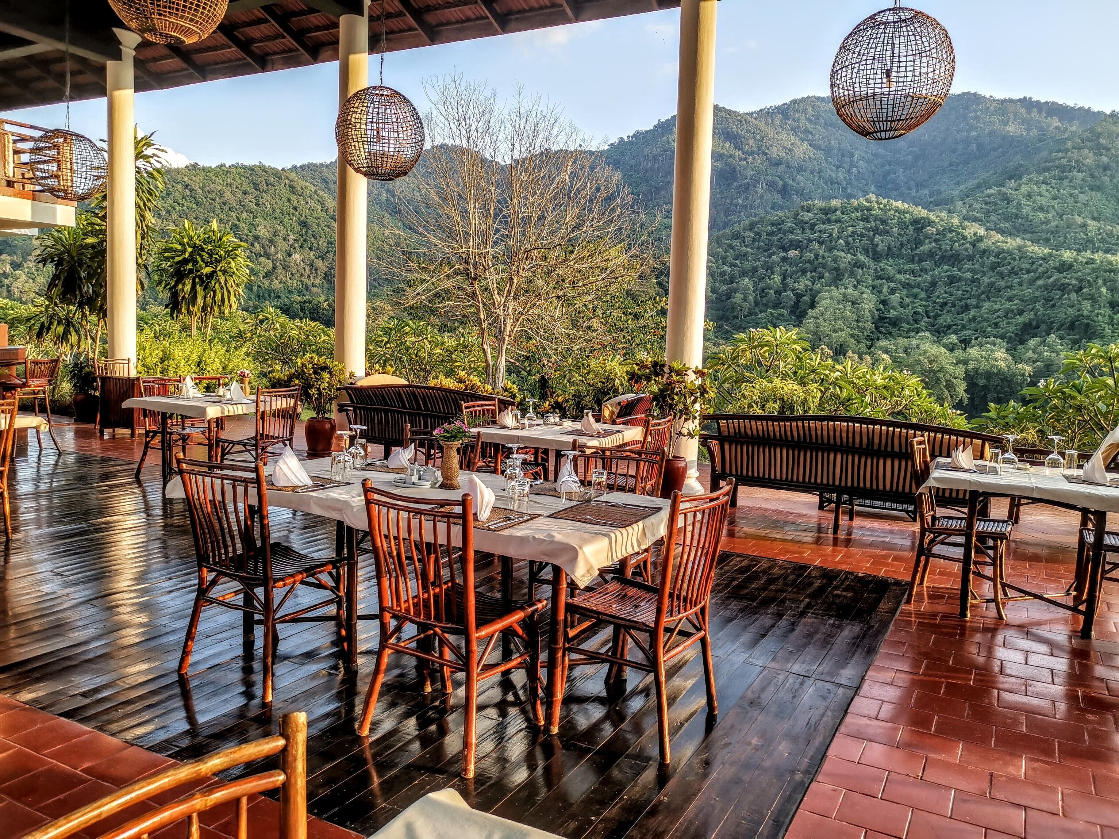 An outdoor restaurant with tables and chairs, surrounded by lush greenery and mountains.