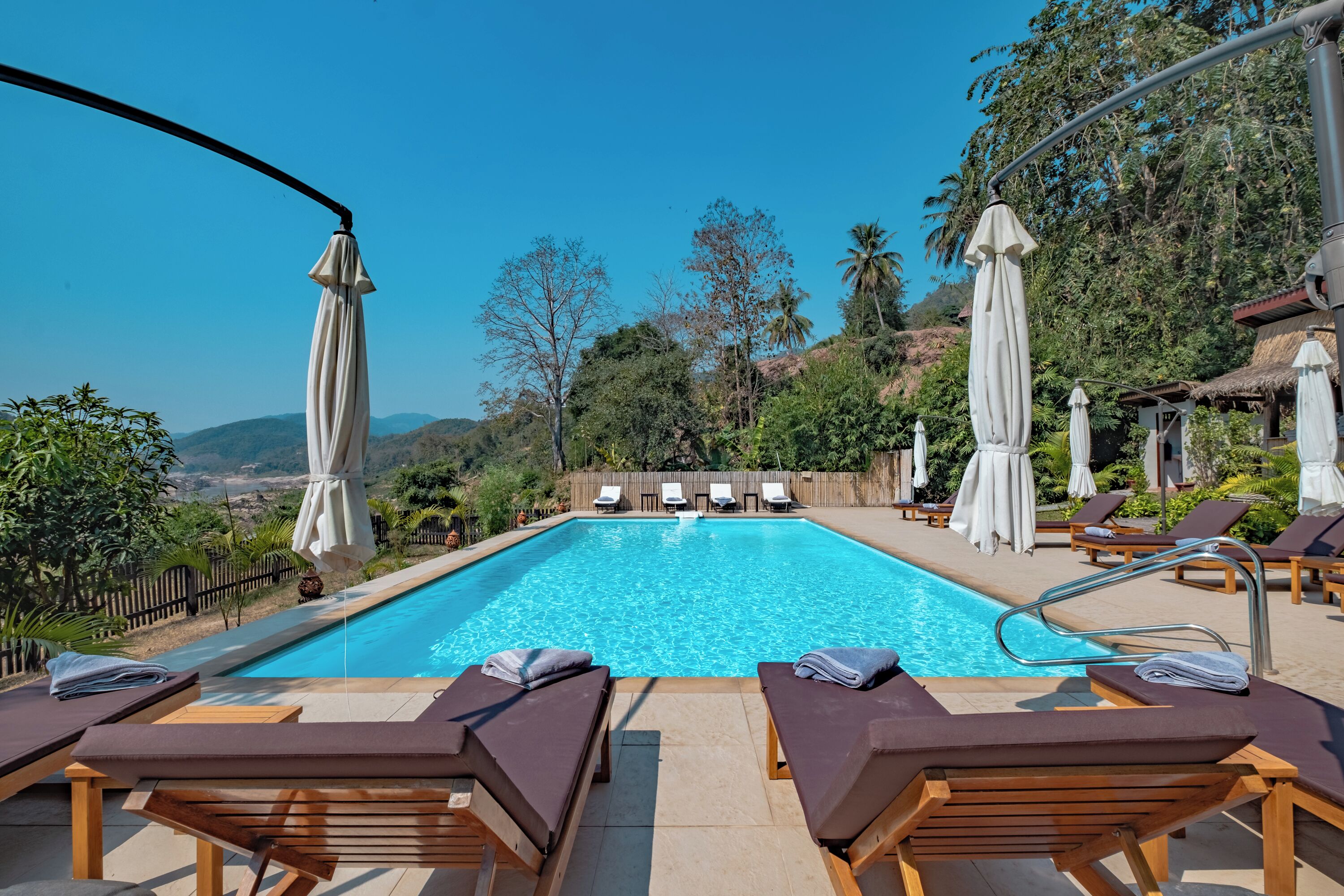 Zona junto a la piscina con sillones de descanso y sombrillas, rodeada de vegetación y colinas.