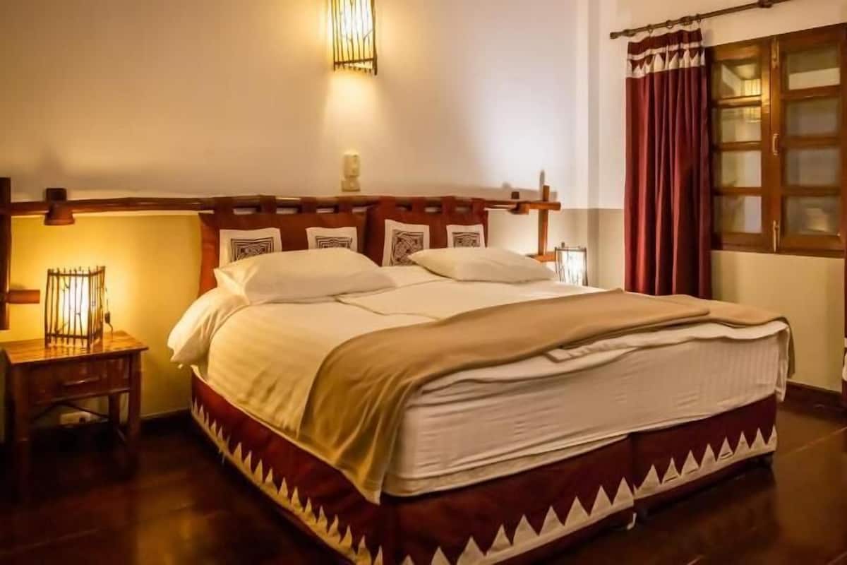 A neatly made bed with a wooden headboard, a bedside table with a lamp, and a window with curtains.