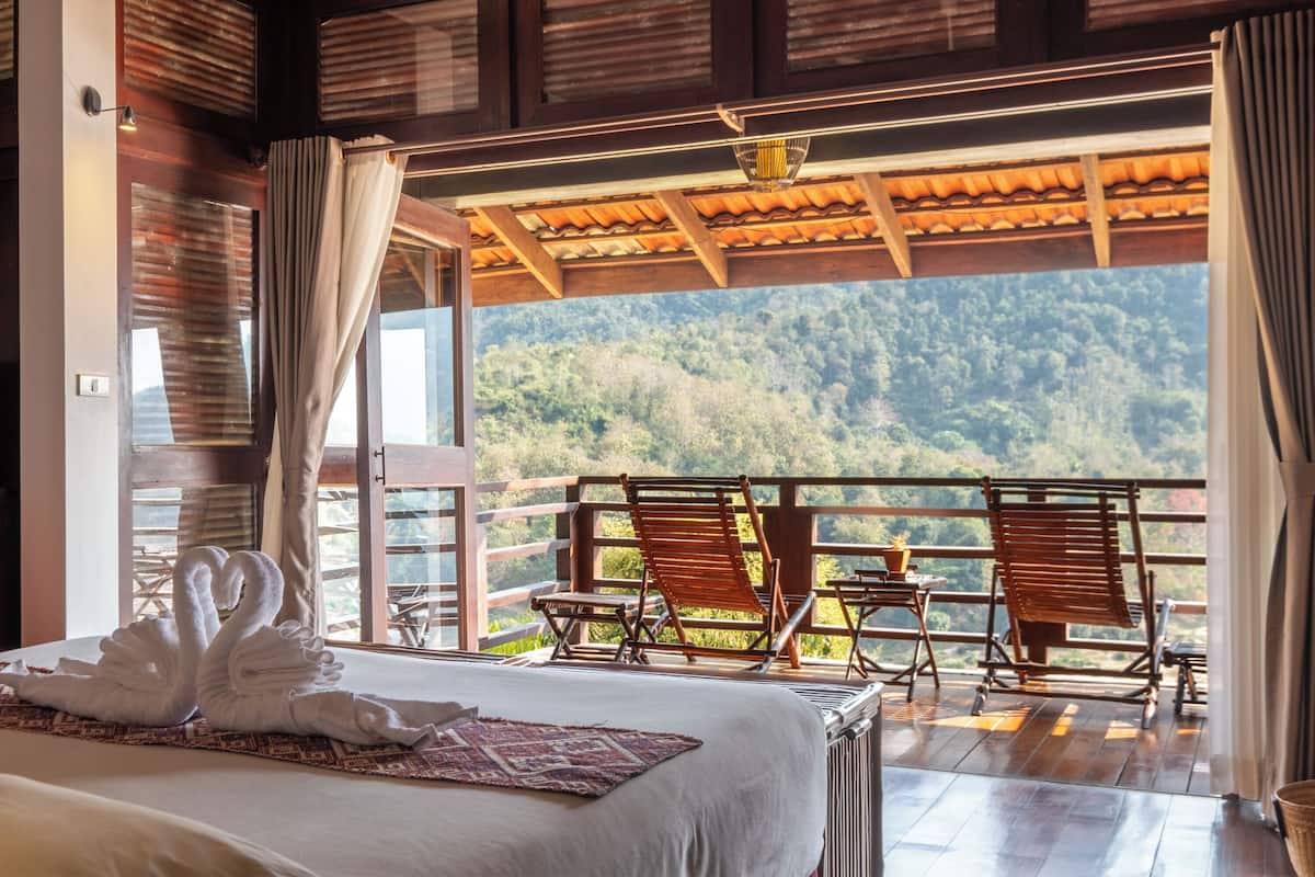 A bedroom with a bed, two wooden chairs, and a balcony with a view of a forested mountain.
