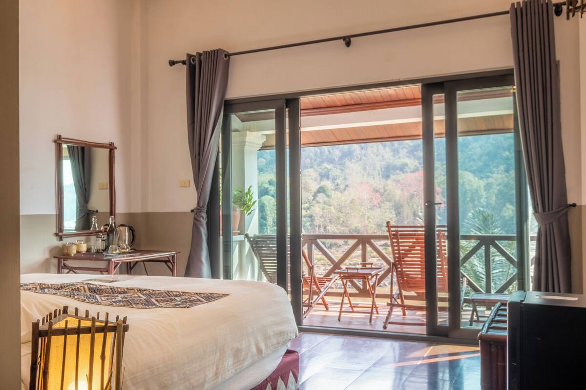 A bedroom with a large bed, a wooden chair, a desk with a mirror, and a balcony with a view of the forest.