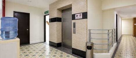 Pasillo de hotel con dispensador de agua, ascensor, contenedor de basura y suelo estampado.