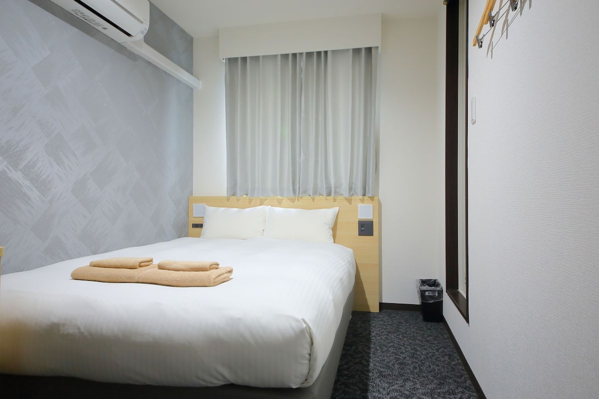 Una habitación de hotel con una cama, una ventana con cortinas, un aire acondicionado instalado en la pared, un pequeño cubo de basura y un interruptor de luz en la pared.
