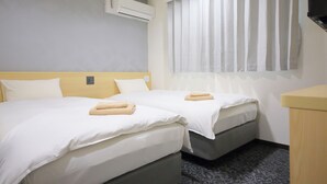 A hotel room with two beds, a window with curtains, an air conditioning unit, and a small wooden desk. - Shin-Imamiya Hotel (Osaka)