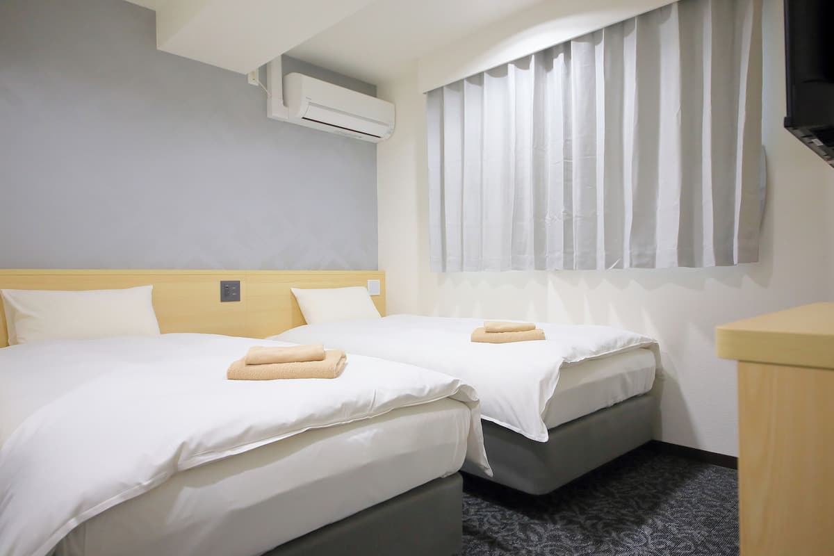 Habitación de hotel con dos camas, una ventana con cortinas, un equipo de aire acondicionado y un pequeño escritorio de madera.