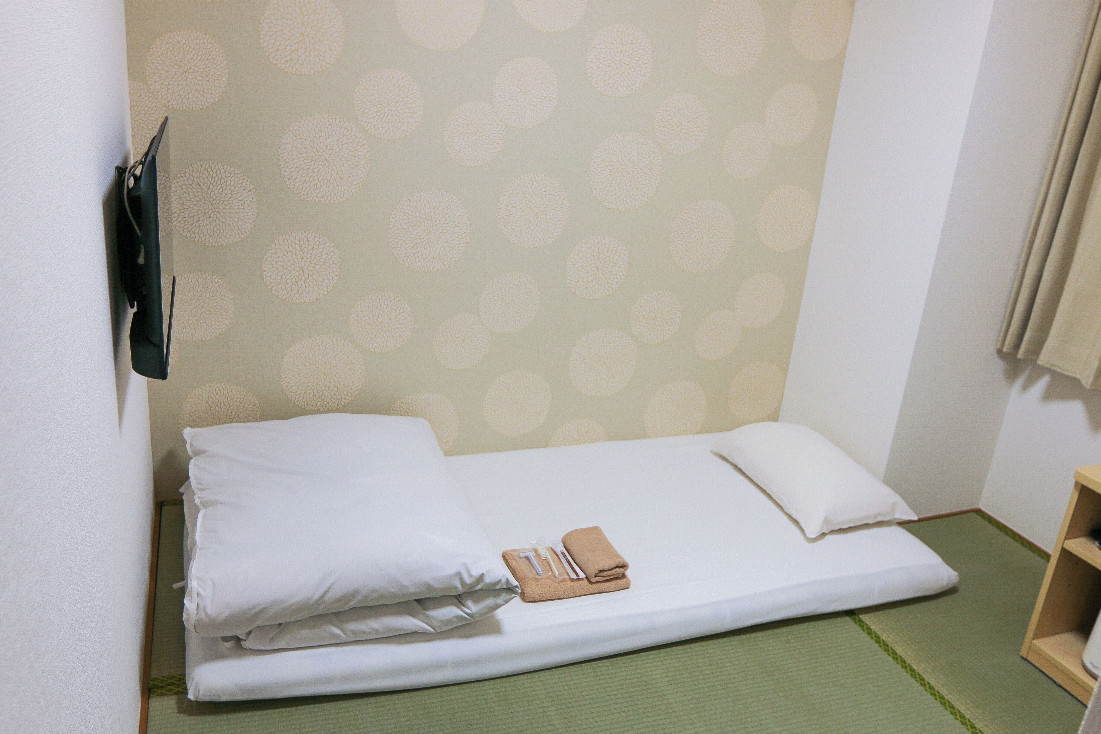 A small, single-bed room with a green tatami mat floor, white bedding, and a wall with a circular pattern.